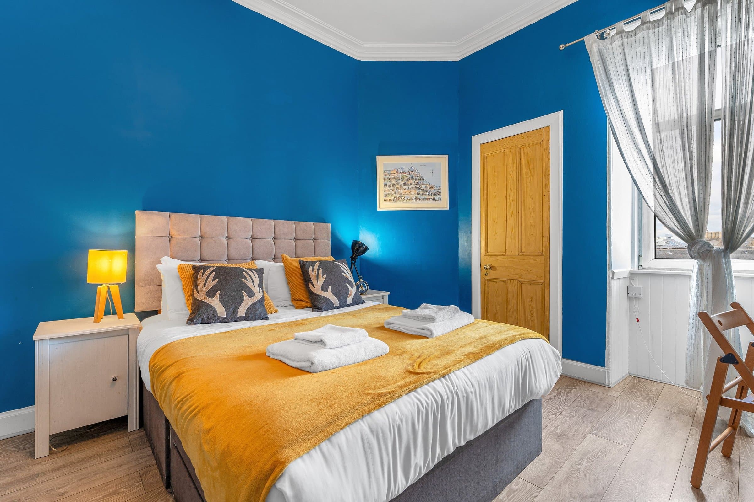 The bedroom, painted deep blue, with a king-size bed under a mustard throw and a tall sash window