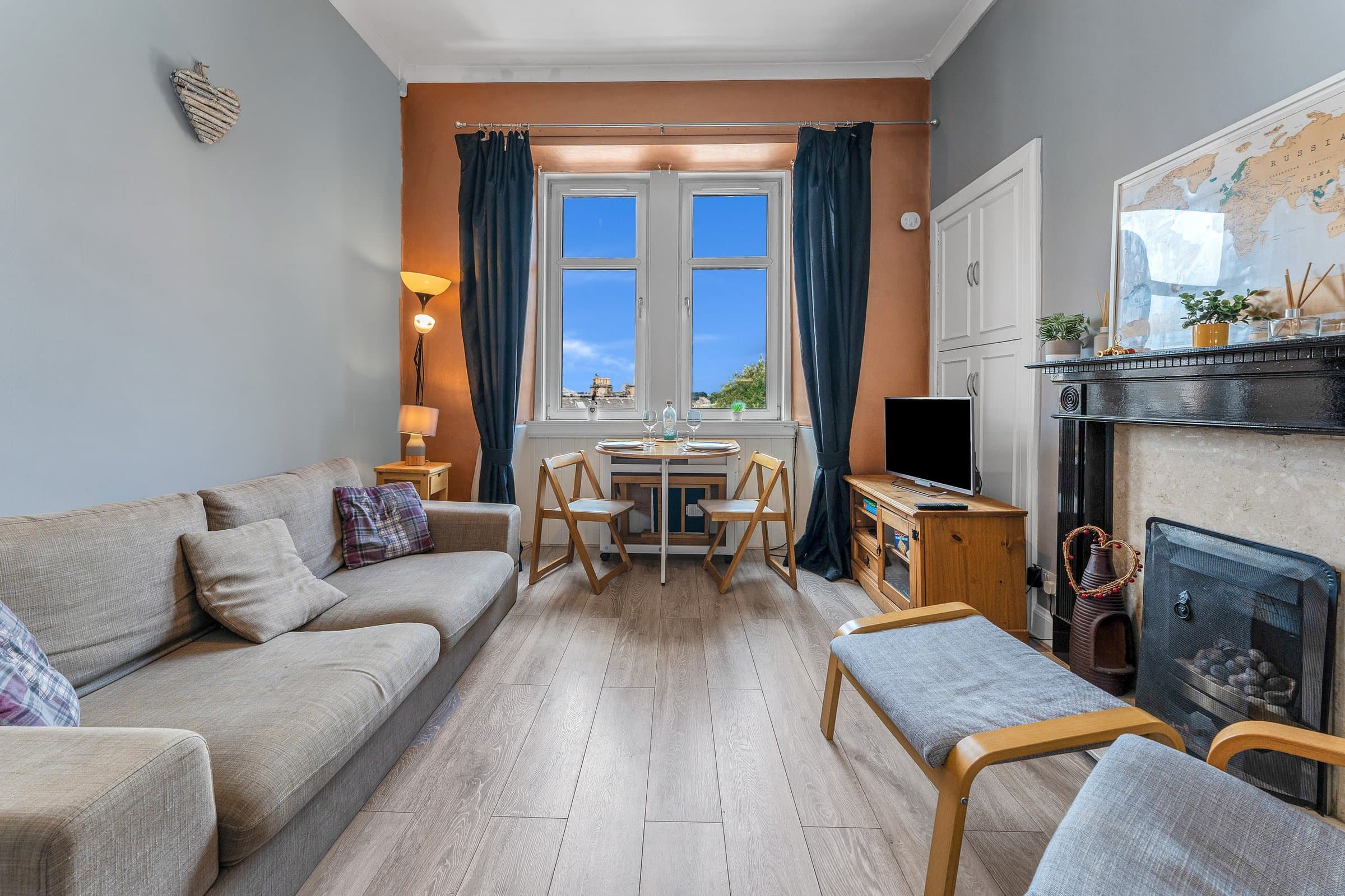 The living room, with a grey sofa and armchair, a black cast-iron fireplace and a small dining table in the bay window looking out over the city